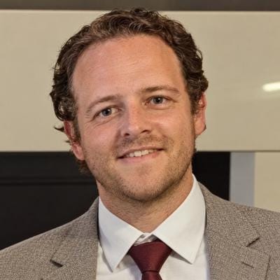 James, founder of UkMoneySheets, smiling in a grey jacket, white shirt and maroon tie.